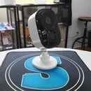 A white security camera mounted on a white stand, placed on a SpyCentre desk mat with a blue graphic, in a retail or office environment.